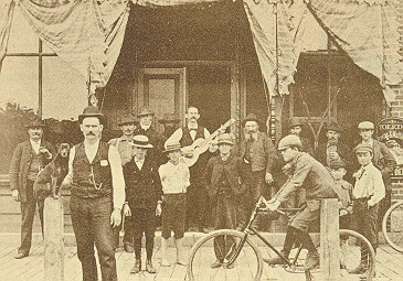 A group of men and boys gather in front of a building. One is on a bike, another holds a guitar, and a small dog is sitting on a wooden pole.