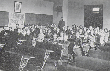 A black and white photo of children sitting at their desks and looking towards the camera while their teacher looks on from the back of the room.